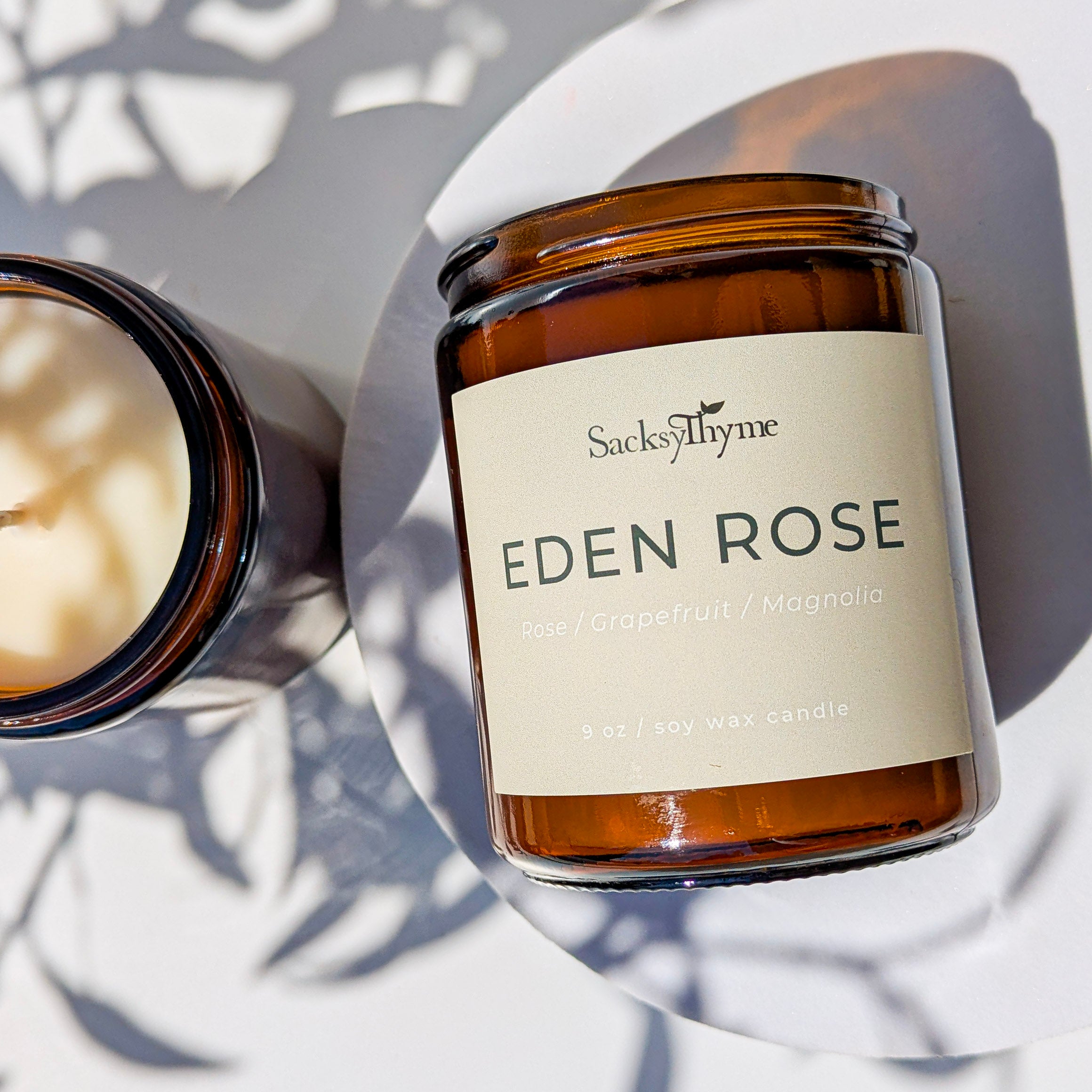 A glass jar candle with the label 'EDEN ROSE' written on it, alongside a partially burned candle, all placed on a surface with a shadow of a plant leaf.