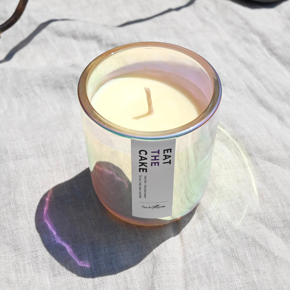 An iridescent glass candle jar filled with cream-colored wax, featuring a white label that reads 'EAT THE CAKE' in black text. The candle is placed on a light gray linen fabric under bright natural sunlight, casting a soft shadow.
