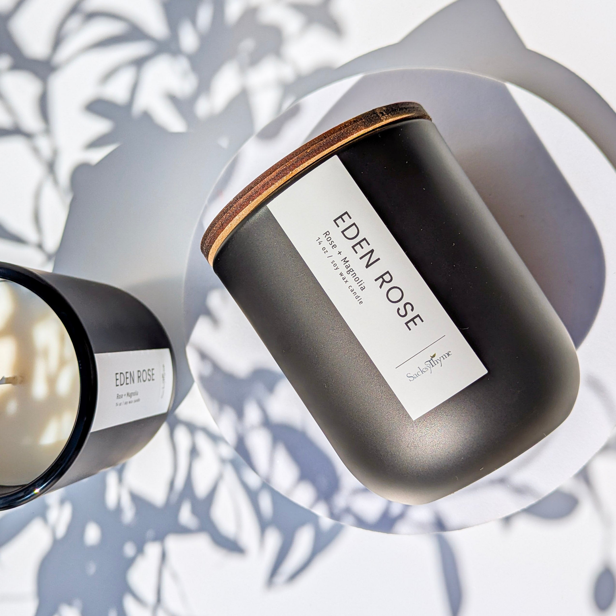 A black glass jar candle with a white label reading 'EDEN ROSE', set on a surface with a shadow of a plant leaf.